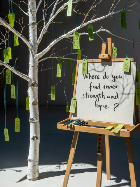 AI generated white birch tree with no leaves stands next to an easel that has a question written on it: "Where do you find inner strength and hope?" Green tags with answers on them hang from the tree.